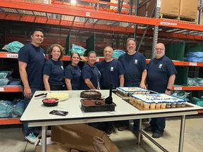 Six people in matching shirts standing behind a table with cakes in a warehouse setting with shelves and boxes.