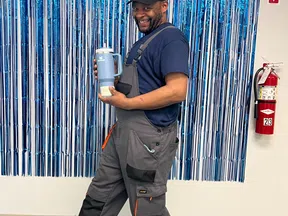Man in gray overalls and cap holding a blue tumbler posing in front of blue fringe backdrop with Cheers to 10 Years sign.