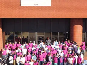 Large diverse group of NOVO Health Services employees posed outside brick building entrance wearing pink and white shirts.