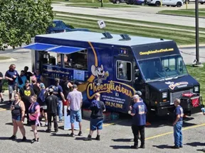 Crowd of people lined up at blue Cheezylicious food truck in sunny parking lot with grass and cars in background