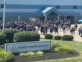 Group of people gathered outside NOVO Health Services building on a sunny day, with landscaping and service vehicles nearby