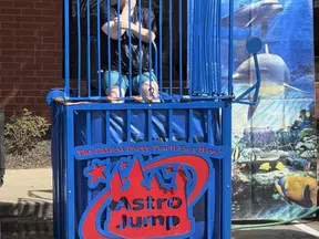 Person sitting in a blue dunk tank cage with Astro Jump logo and aquatic-themed backdrop outdoors.