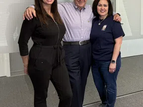 Three smiling adults standing indoors with arms around each other in casual and business attire.