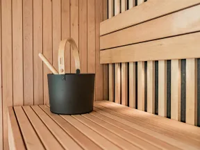 Black sauna bucket with wooden handle and ladle inside a light wooden sauna room interior.