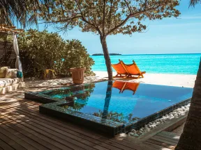 Infinity pool overlooking tropical beach with turquoise water and two orange lounge chairs under tree shade.