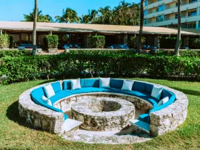 Outdoor circular stone seating area with turquoise cushions and white pillows under palm trees on a sunny day.