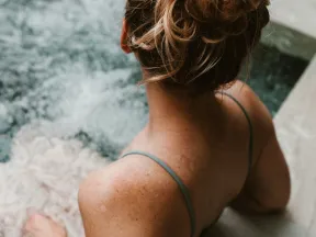 Woman with blonde hair bun relaxing in a hot tub with clear water and bubbling jets, viewed from behind.