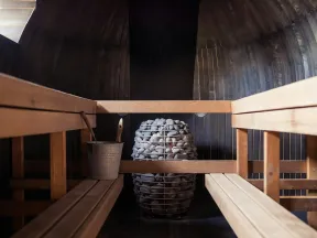 Interior of wooden sauna room with benches, metal bucket, ladle, and stone heater in the center.