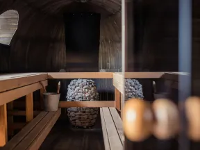 Interior of a wooden sauna with benches, a bucket, ladle, and a stone heater in a cozy setting.