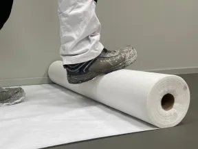 Person wearing work shoes and white pants rolling out a large white protective floor cover on gray surface indoors.