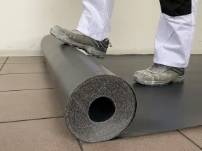 Person in workwear unrolling black flooring material on tiled floor with foot on the roll.