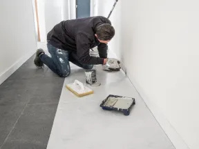 Person kneeling in a hallway painting baseboards white with painting tools scattered around.