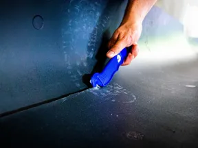 Hand using a blue utility knife to cut thick black foam or insulation material on a flat surface.