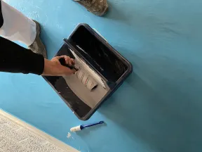 Person dipping a paint roller into white paint on a blue floor with a small brush nearby.