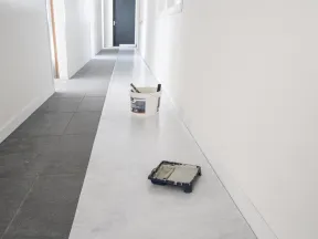 Long hallway with white walls, grey tiled floor, paint bucket, and tray with roller for renovation work.