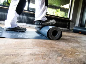 Worker installing black protective floor lining on wooden flooring inside a modern room with large windows.