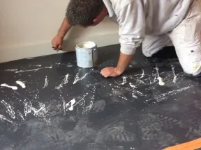 Painter kneeling on black covered floor applying white paint to wall baseboard with a brush.