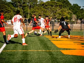 Soccer players in red and white jerseys competing near the goal on a sunny outdoor field with green grass.