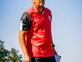 Coach in a red and black sports outfit stands outdoors holding a whistle and a clipboard under clear blue sky.