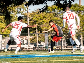 Young soccer player in red and black challenges two defenders in white jerseys on green field with trees.