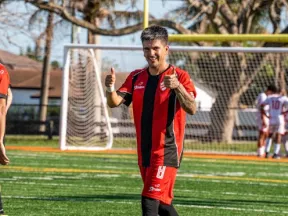 Soccer player in red and black uniform giving thumbs up on green artificial turf field near goalpost.