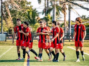 Soccer team in red and black jerseys walking and talking on a sunny field with palm trees in the background.