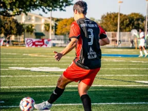 Soccer player in red and black uniform kicking a ball on a sunny outdoor field with trees and buildings in the background.