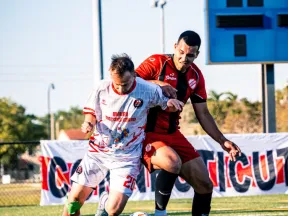 Two soccer players contesting for the ball on a sunny field with a scoreboard and banner in the background