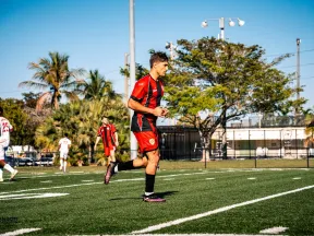 Soccer players in red and white uniforms on a sunny field with palm trees and goalposts in the background.