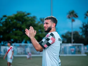 Soccer player in a white and checkered jersey clapping on a field with blurred background at dusk.