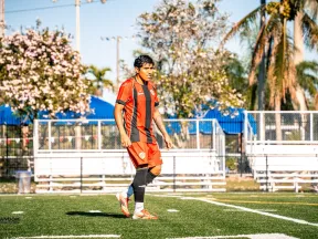 Soccer player in red and black uniform walking on field with palm trees and benches in background under clear sky