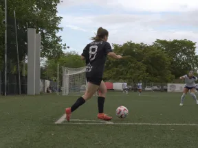Female soccer player in black jersey preparing to kick ball near goal on grassy field during game.
