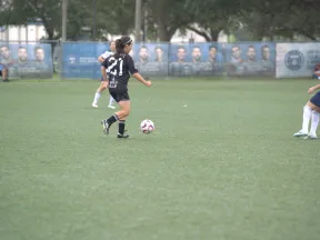 Female soccer player in black jersey controlling the ball on a green field against opponents in white jerseys.