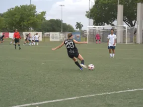 Female soccer player in black jersey kicking ball on outdoor field during game with opponents and referee.