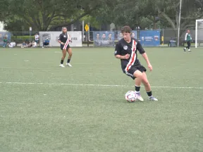 Youth soccer players in black jerseys with red and white stripes playing on a green field during a daytime match.