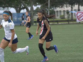 Female soccer players in action on a green field during a daytime match with trees and a fence in the background