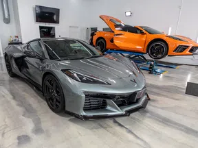 Gray Corvette sports car parked in a garage with an orange Corvette on a lift with its hood open.