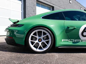 Green Porsche sports car side view showing rear wheel, aerodynamic spoiler, and partial brand logo on sunny day.