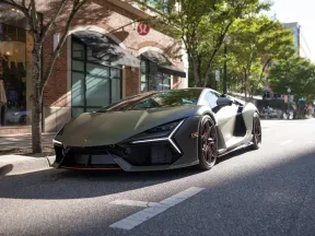 Matte green Lamborghini sports car parked on a sunlit urban street with trees and shops in the background