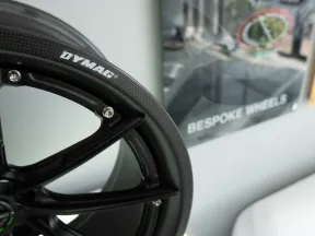 Close-up of a black Dymag carbon fiber alloy wheel with a bespoke wheels poster in the background