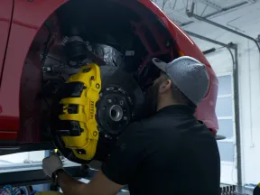 Mechanic inspecting a red car's yellow brake caliper in a garage with raised lift and tools nearby