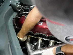Person repairing a red engine with shiny exhaust pipes inside a car using gloved hands.