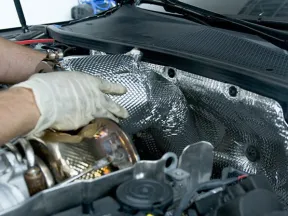Technician wearing gloves installs heat shield inside a car engine compartment for thermal protection.