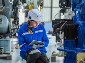 Female engineer in blue safety gear inspecting machinery in an industrial factory setting using a tablet.