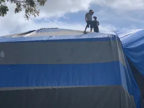 Two people standing on a large blue and gray tarp covering a roof under a cloudy sky