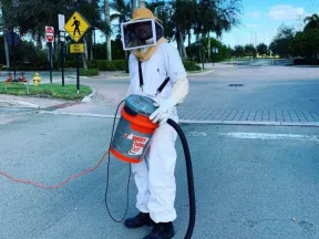 Person in protective beekeeper suit holding a homemade bee vacuum on an urban street.