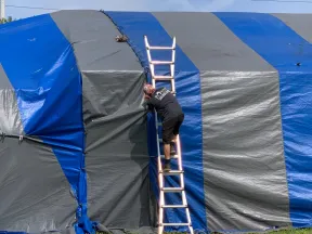 Person climbing a ladder to adjust a large blue and gray striped outdoor tent under a partly cloudy sky.
