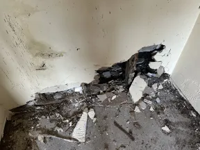 Damaged corner of room with broken drywall, exposed wood, and debris on floor indicating structural decay.