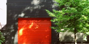 Modern black garage with vibrant red door next to green leafy tree on sunny day