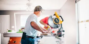 Two men cutting wood with a miter saw inside a bright, white room under renovation.